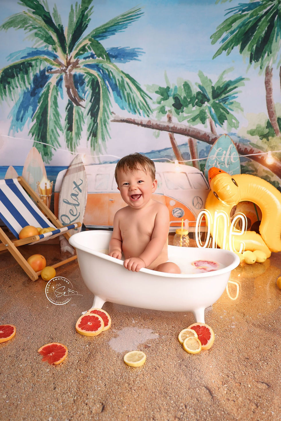 Bébé souriant assis dans une baignoire blanche sur une plage décorée, avec palmiers, rondelles d’agrumes et le mot one.