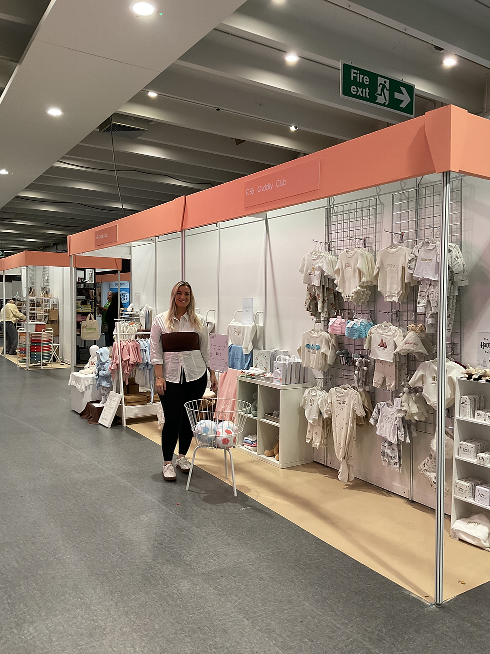 The image shows Catherine-Emma, the owner and creator of Cuddily Club with the stall at The Baby And Toddler Show in Sandown, London in September 2025. You can see many different handmade baby items like baby pyjams, baby clothing sets, knitted baby blankets, hand crochet rattles, baby books.