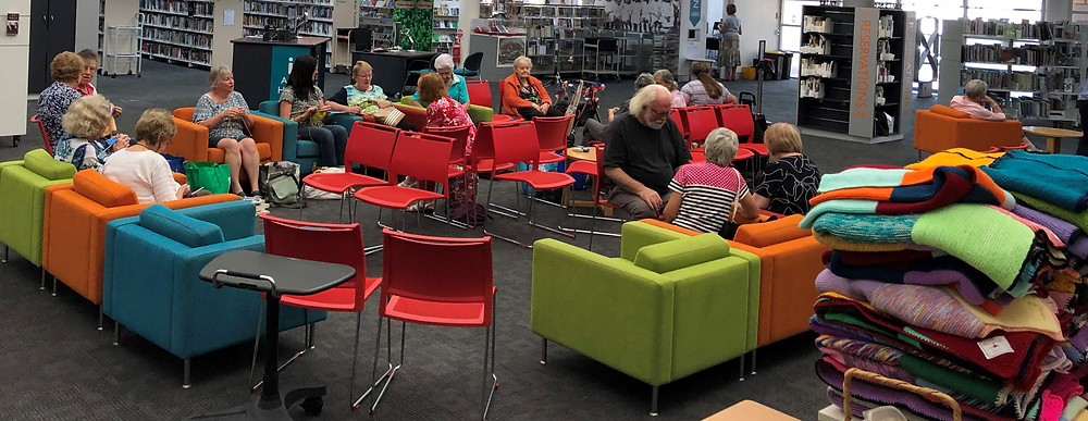 A group of around 15 people are seating in colourful armchairs, knitting and chatting.