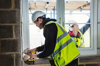A construction worker in a white hard hat and yellow safety vest measures wood