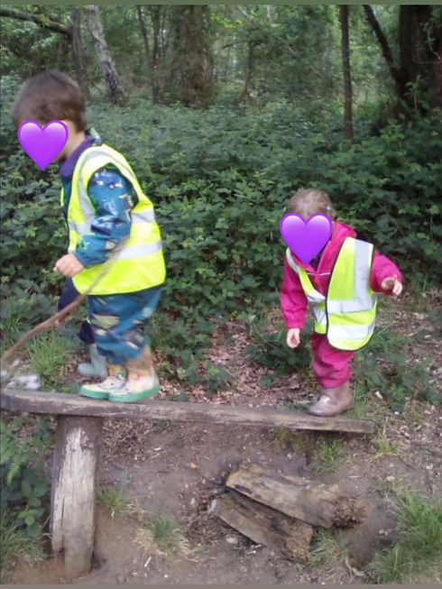 Two young children in high visibility vests playing outdoors near trees and wooden logs