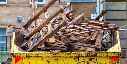 A yellow skip bin filled with irregularly stacked, weathered wooden planks against a blurred urban backdrop