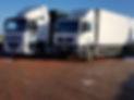 Two white delivery trucks parked on a dirt lot under clear blue skies