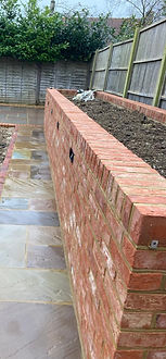 A newly constructed garden features a red brick raised bed on the right, a tiled path in the center