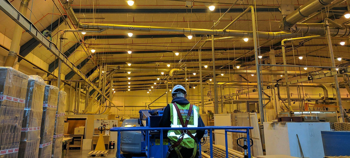 A worker in safety gear and a hard hat stands on a lift inside an illuminated industrial warehouse