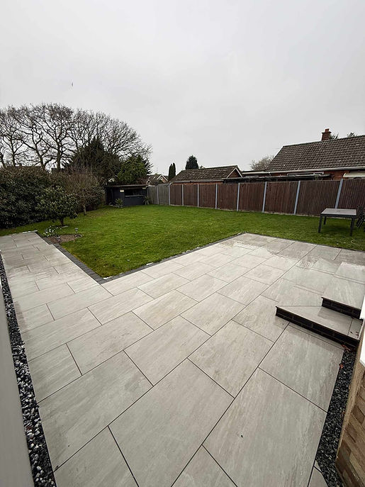 L-shaped light grey porcelain patio with black gravel borders beside a green garden lawn