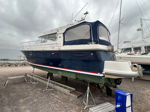 Boat on land with a dark blue hull and white cabin, supported by stands in a boatyard