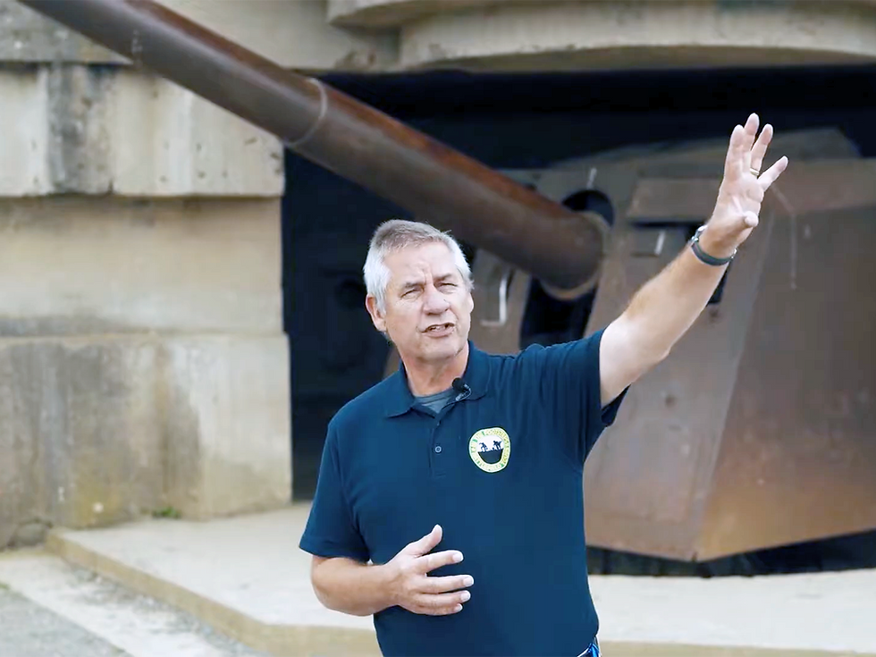 A man in a polo shirt gestures while standing in front of a large military gun or turret.