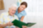 An elderly man smiles while reading a large green book with a caring woman