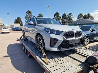 A silver SUV with a bold front grille is secured on a flatbed trailer in a parking lot