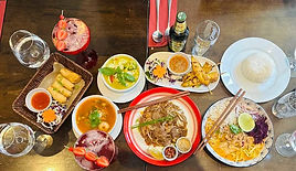 A variety of dishes arranged on two tables, including spring rolls, rice, a bowl of soup, a plate of noodles, and various sauces and drinks, with decorative garnishes.
