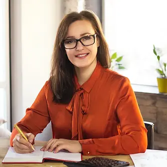 Smiling woman in glasses and an orange blouse writing in a notebook at a desk
