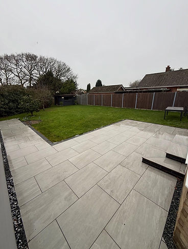 Backy view featuring a tiled patio and green lawn, bordered by bushes and a wooden fence