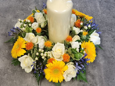 A decorative arrangement with a large white candle surrounded by yellow gerbera daisies