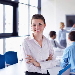 Smiling woman in a white shirt stands confidently with arms crossed in a bright office