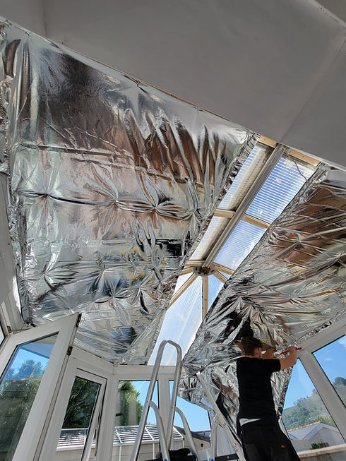 A person stands on a ladder, attaching reflective foil insulation to a greenhouse ceiling