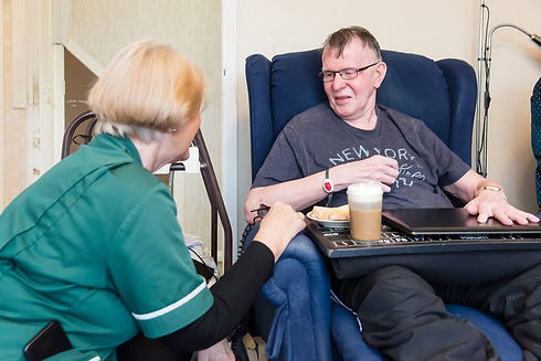 An elderly man smiles in a blue armchair, holding a drink on a tray