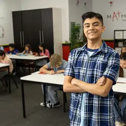 Teenage boy in a checked shirt standing proudly with crossed arms in a busy classroom