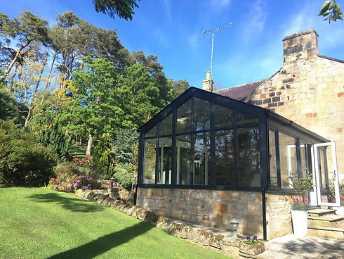 A sunlit glass conservatory extends from a stone house surrounded by lush greenery and colorful flowers
