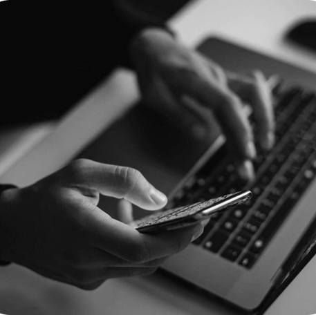 A close-up black and white image of hands multitasking with a smartphone and a laptop