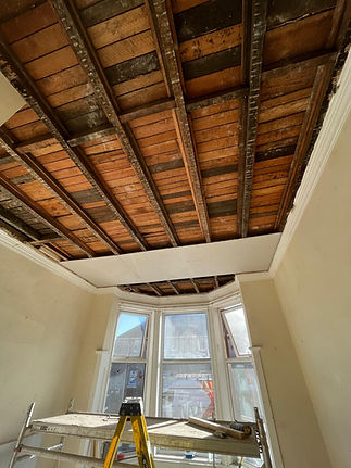 Exposed wooden beams in a ceiling under renovation, with sunlight streaming through bay windows