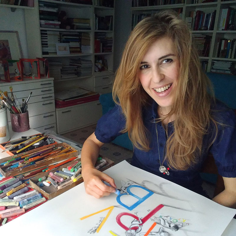 Smiling Author Eva Montanaridraws a colorful sketch at a desk covered with pastels in a cozy art studio with bookshelves.
