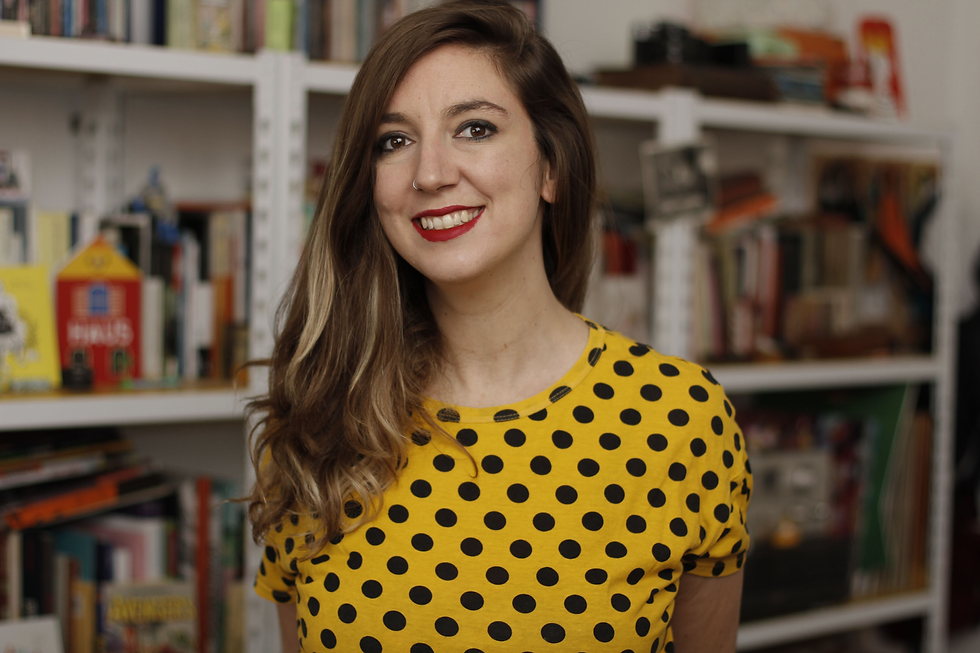 Smiling Mariana Ruiz in a yellow polka-dot shirt stands before bookshelves in a cozy room.