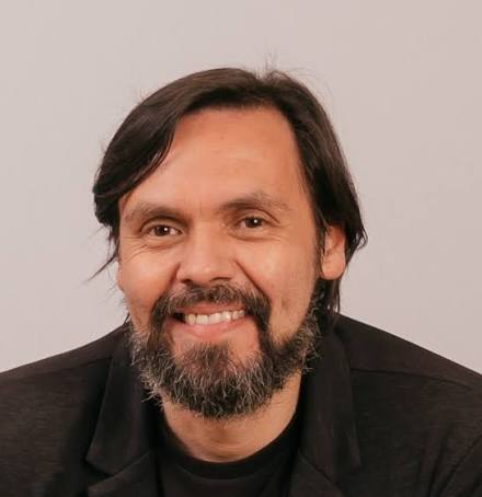 Artist Gerald Espinoza with dark hair in a black jacket, posed against a plain light gray background.