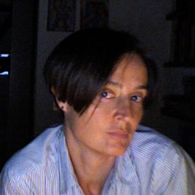 Portrait of author Giovanna Zoboli with short dark hair in a striped shirt, looking at the camera in a dim room with a serious expression.