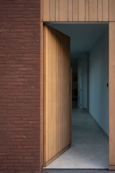 De houten voordeur van deze prachtige houtskeletbouw woning in Tielrode (Temse) straalt warmte en karakter uit. Volledig bekleed met hout, reikt de deur tot aan de dakrand en vormt zo een naadloos geheel met het architecturale ontwerp. De woning, met zijn centrale patio als kloppend hart, nodigt de natuur binnen en creëert een harmonieuze verbinding tussen binnen- en buitenruimte. Bij het betreden van de woning onthult de voordeur een direct zicht op de tuin via de patio, waar het spel van licht en schaduw een serene sfeer creëert. Het gebruik van hout weerspiegelt niet alleen vakmanschap, maar draagt ook bij aan de warmte en gezelligheid van de entree. Een boeiend staaltje architectuur in een rustige, lichtrijke omgeving, vastgelegd op beeld door Rebecca Tanghe Fotografie.