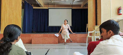 A facilitator seated on stage in front of a projector screen, speaking to a small group of participants during an introspective storytelling and reflection workshop.