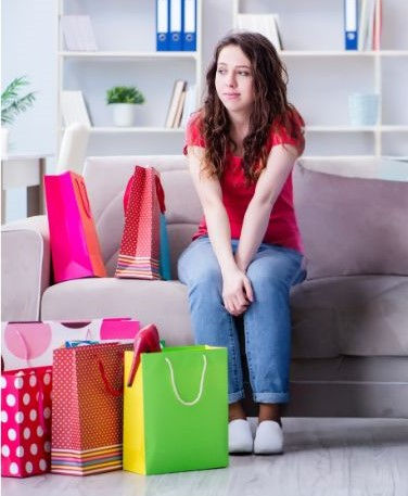 Eine junge Frau sitzt auf einer Couch umringt von ihren Einkäufen und hat ein schlechtes Gewissen