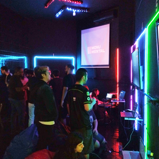 Pessoas estão reunidas em pé em uma sala escura com luzes azuis e verdes enquanto conversam e jogam videogames