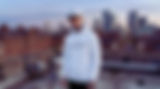 A tattooed male model stands alone on a UK city rooftop at dusk, wearing a White Blazed We