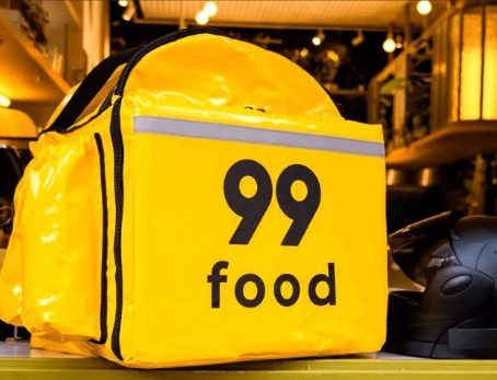 Mochila térmica amarela da 99Food com o logotipo “99 food”, apoiada sobre um balcão, ao lado de um capacete de entregador.