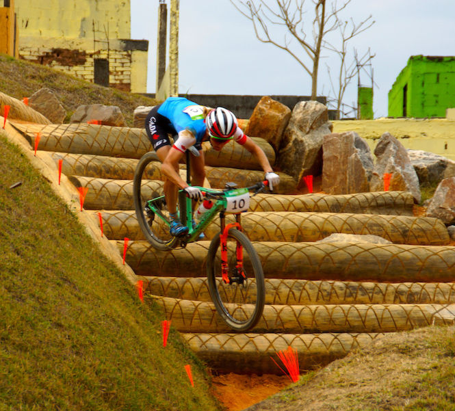 Cyclist in a blue jersey descends a rocky, log-paved path with vibrant green and yellow buildings in the background. Number 10 is visible.