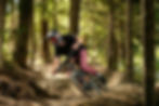 Mountain biker in helmet rides through a forest trail, leaning into a turn. Sunlight filters through trees, creating a dynamic, energetic vibe.