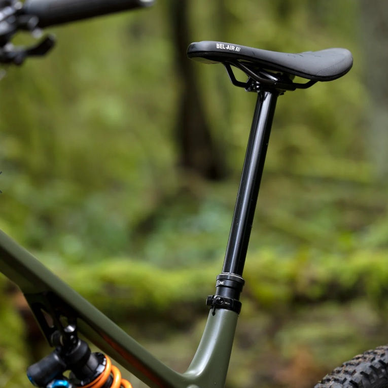 Close-up of a bike seat labeled "Bel-Air III" in a forest setting, with a green frame and coil shock. Background is lush and green.