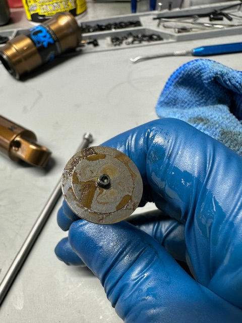 Blue-gloved hand holds a rusty metal disk on a workbench, with tools and a blue cloth in the background.
