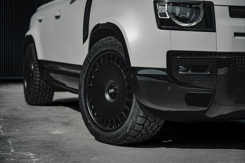 Modified white Land Rover Defender D250 with black roof and aggressive black off-road wheels, parked in front of a modern industrial metal wall.