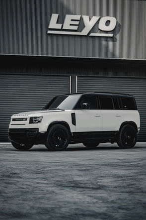 Modified white Land Rover Defender D250 with black roof and aggressive black off-road wheels, parked in front of a modern industrial metal wall.