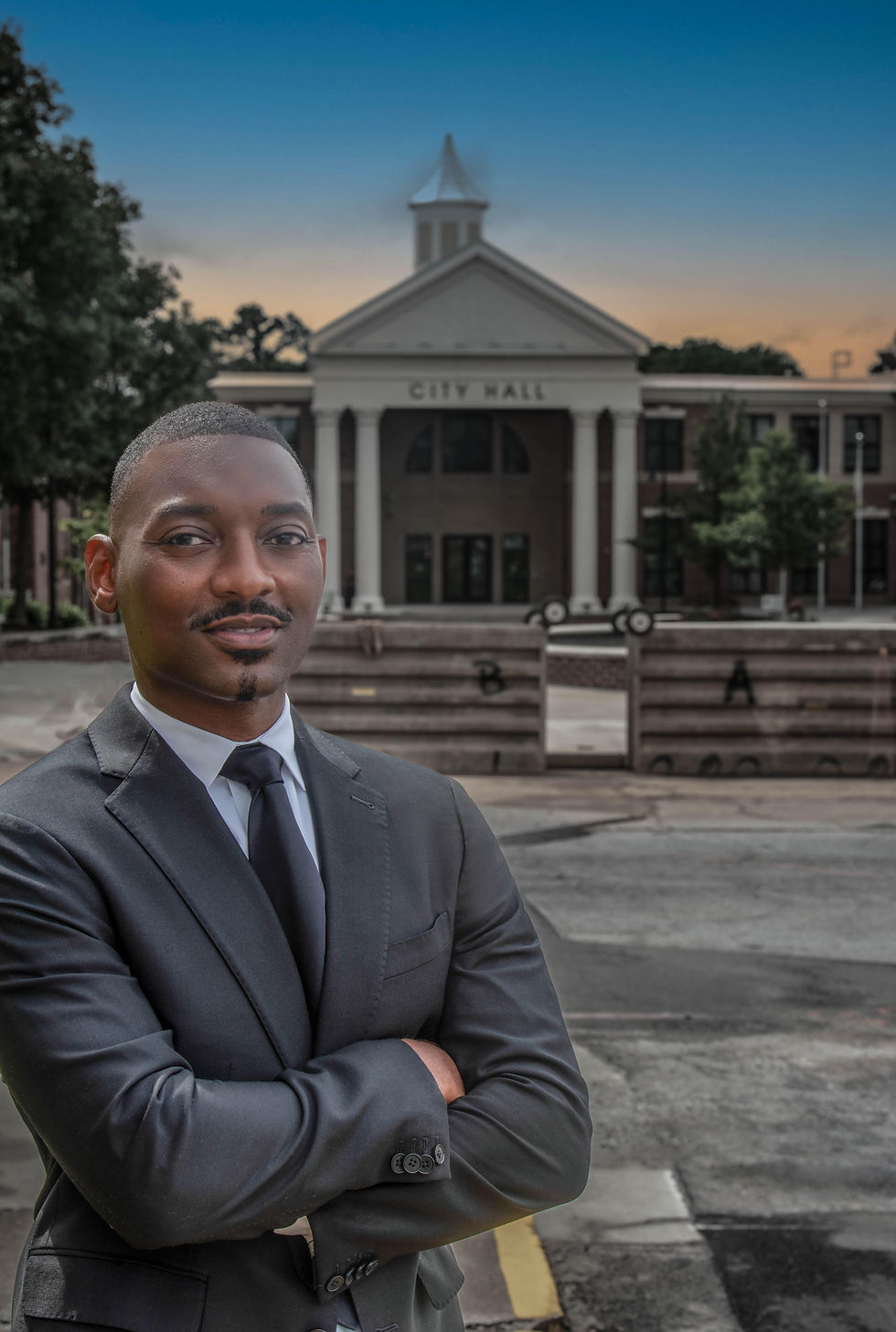 Erik Coleman standing in front of East Point City Hall, wearing a black suit, white shirt, black tie, outdoors in daylight