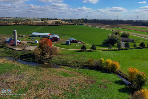 Scenic rural landscape with a farm, fields, and a winding stream under blue sky.