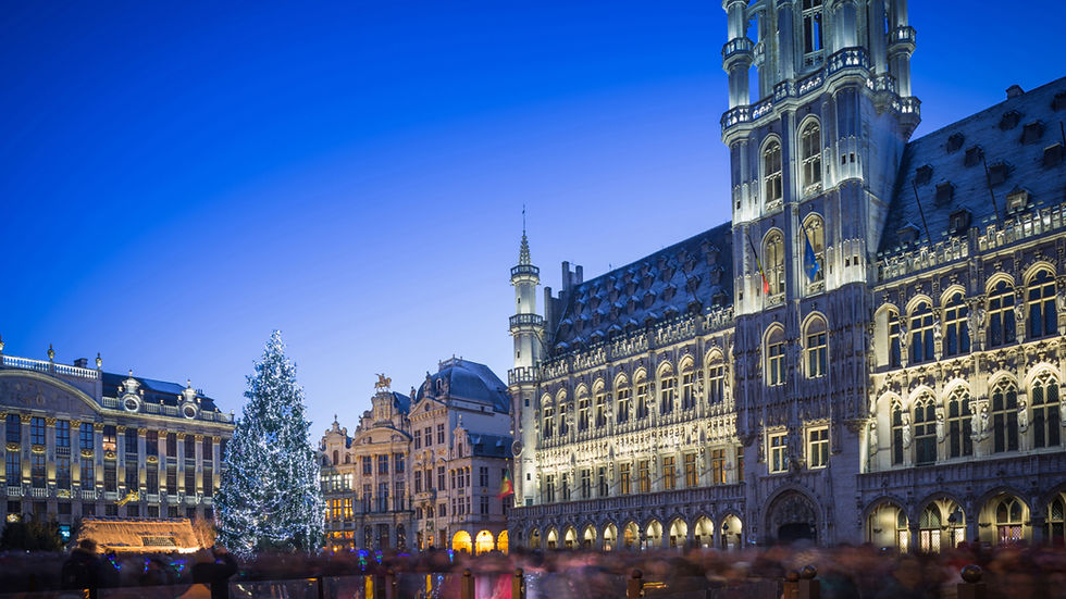 A twilight photograph of the illuminated Grand-Place (Grote Markt) in Brussels, Belgium, as depicted in "Belgium Christmas.jpg". The magnificent, fully lit Brussels Town Hall with its central tower stands prominently on the right, set against a clear, deep blue sky. A large, motion-blurred crowd of people fills the square below, creating a sense of movement. To the left, a large, decorated Christmas tree covered in cool white fairy lights stands next to an illuminated Nativity scene stall. Historic, decorated guildhalls line the square, which is bustling with the blurred crowd and festive lights. The lighting is warm and clear, emphasizing the gothic architecture against the dusk sky.