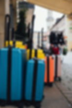 Colorful suitcases lined up on a cobblestone street. Yellow, blue, orange luggage with extended handles beside a shop under a canopy; cityscape blurred.
