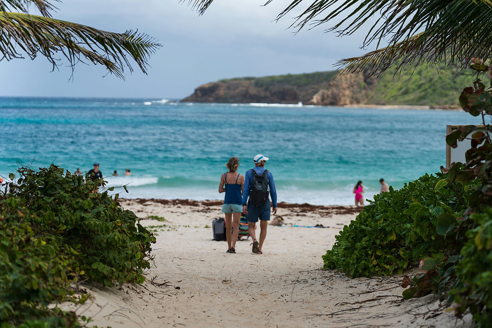 Couple walking on a sandy path toward a beach, surrounded by lush greenery. People swim in turquoise sea under cloudy sky. Relaxed mood.