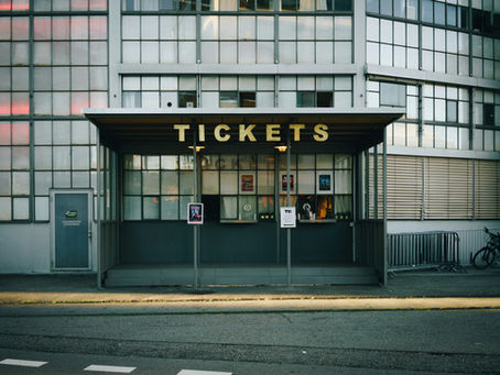Ticket booth with a retro design, large "TICKETS" sign, and posters on the window in front of a modern industrial building facade.