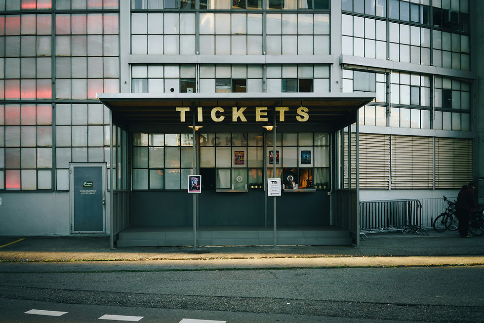 Ticket booth with a retro design, large "TICKETS" sign, and posters on the window in front of a modern industrial building facade.