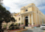 Historic yellow building with "El Convento" sign, arched entrance, and columns. Person walking near plants and a parked white van.
