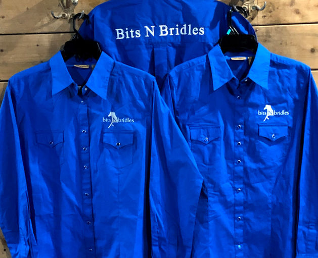 Three blue shirts hanging up on a wooden wall. The two front shirts have the BitsNBridal logo. The back one says BitsNBridles in big white letters.